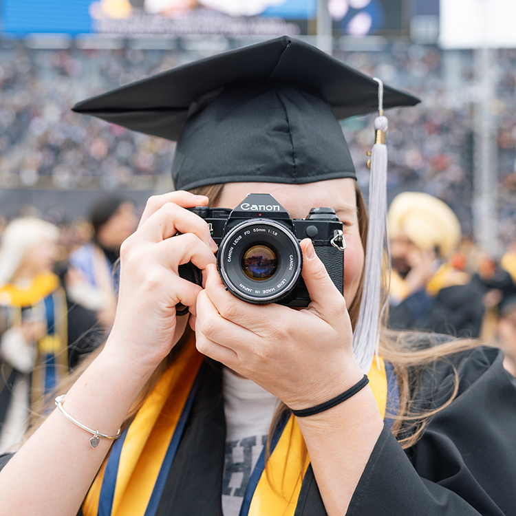 A graduate in a cap, and gown holds a vintage Canon camera up to their eye and looks through the viewfinder at the 2025 Spring Commencement ceremony in the Big House.