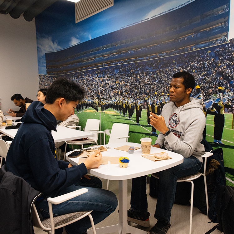 Two students sit at a table and enjoy bagels and coffee in front of a mural of the Michigan Marching Band playing on the field at Michigan Stadium during Welcome Wednesday at the Alumni Center.