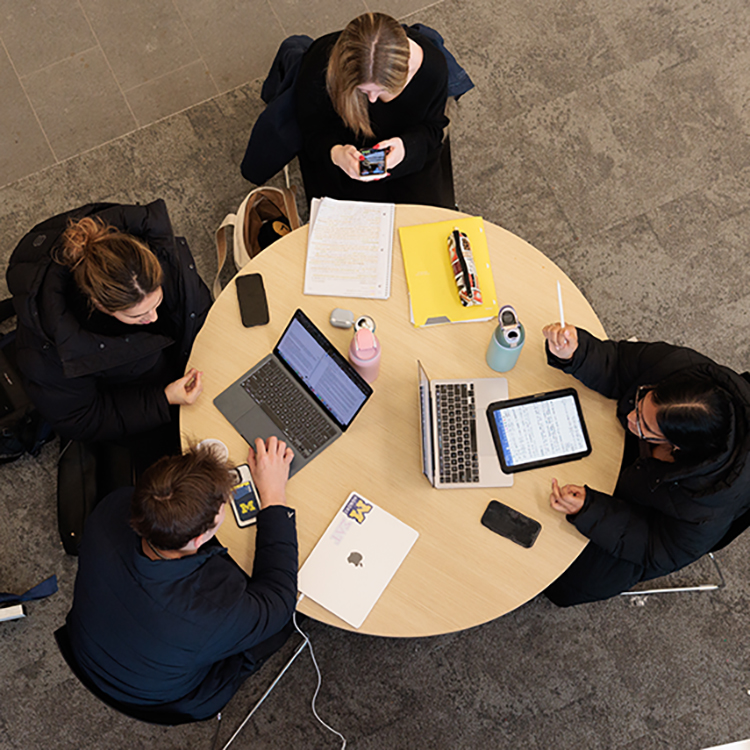 Students study at seats around a table in the LSA building. The tabletop is crowded with notebooks, folders, and laptops.